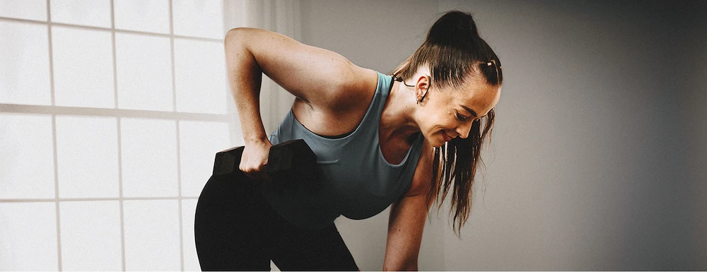 Woman performing bent-over dumbbell row exercise in a well-lit room with large windows.