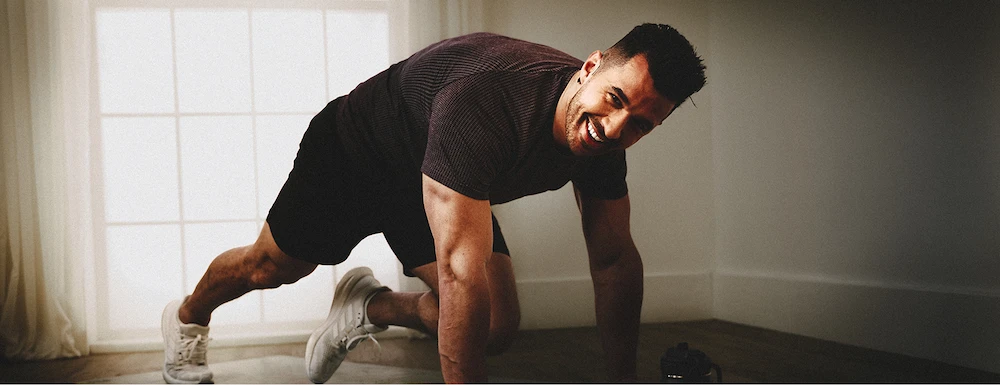 Man smiling while doing a plank exercise in a room with a large window and light curtains.