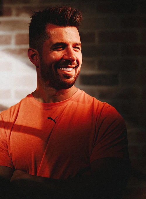 Smiling man in an orange Puma shirt with folded arms standing against a brick wall in warm light.