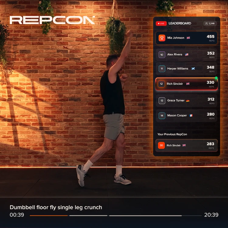 Man in black shirt and gray shorts doing single leg crunch in front of a leaderboard on brick wall.