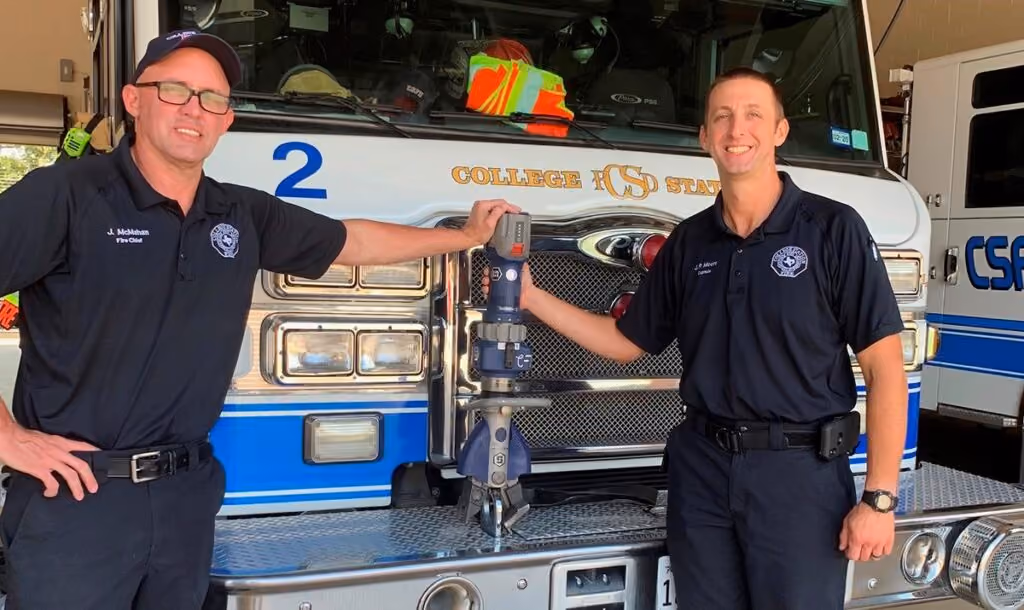 Two fire fighters standing in front of a truck while holding the Jaws of Life