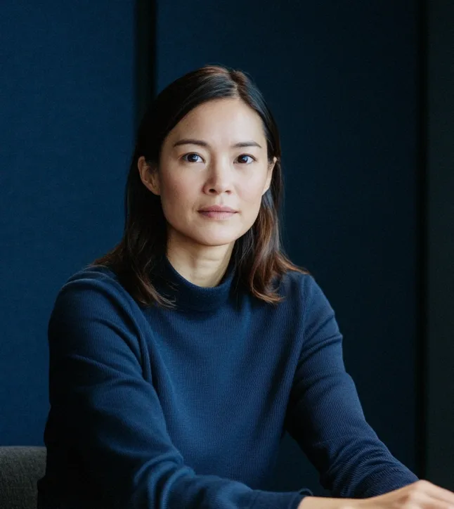Portrait of a woman with dark hair wearing a navy blue sweater against a dark background.