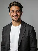 Smiling man with curly hair wearing a black blazer and white t-shirt on a gray background.