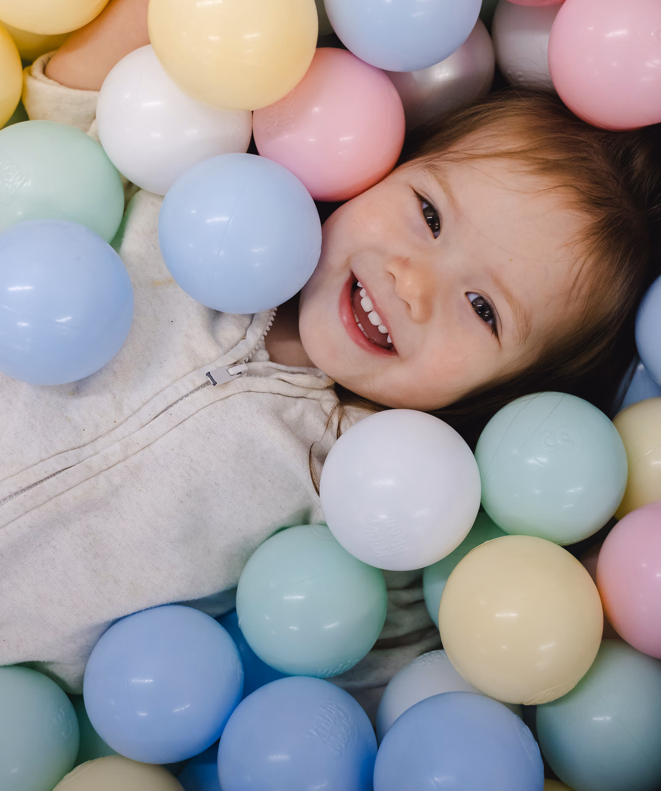 Smiling child lying among pastel-colored plastic balls in a ball pit.