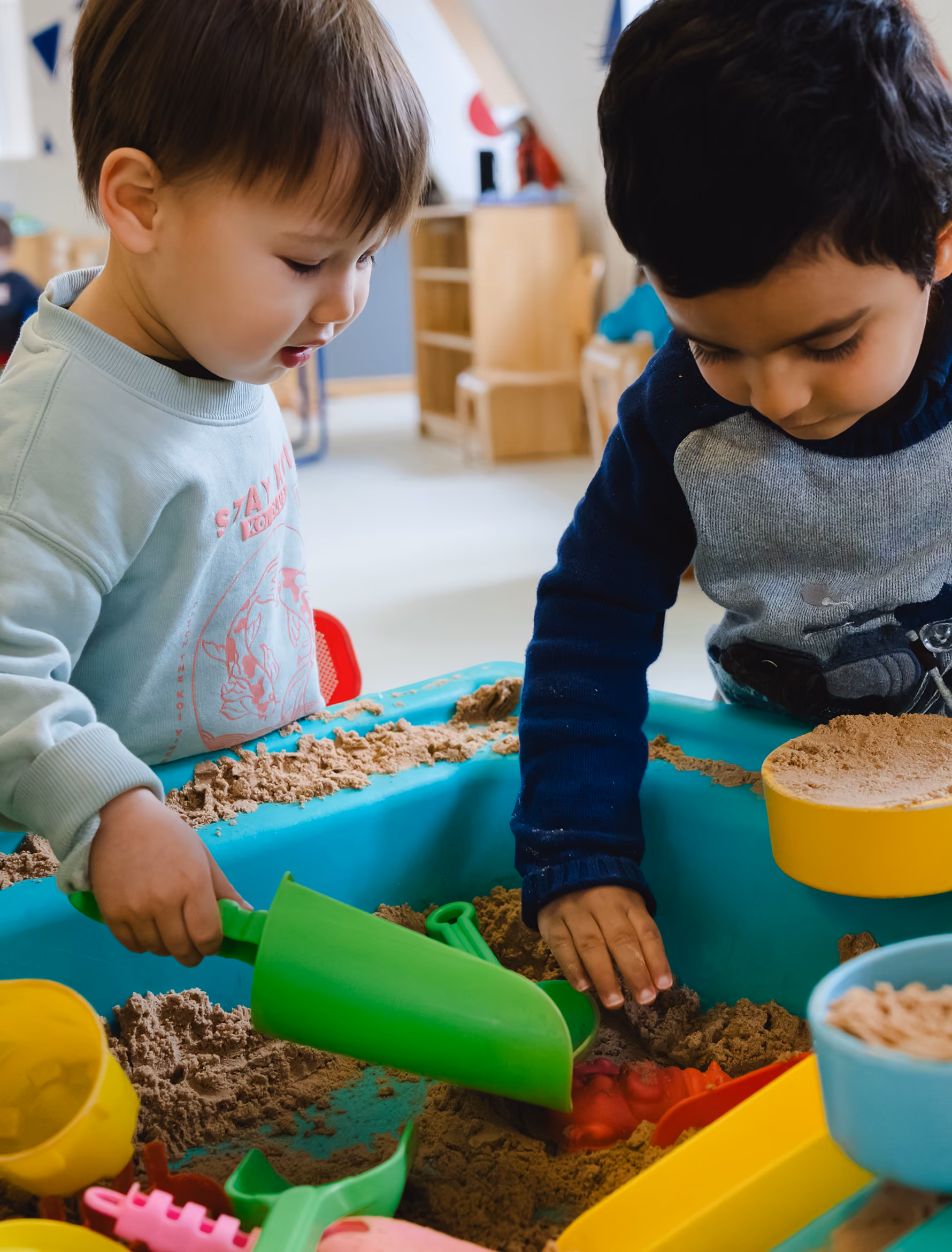 Two young children playing with sand and colorful toys in a blue sandbox indoors.