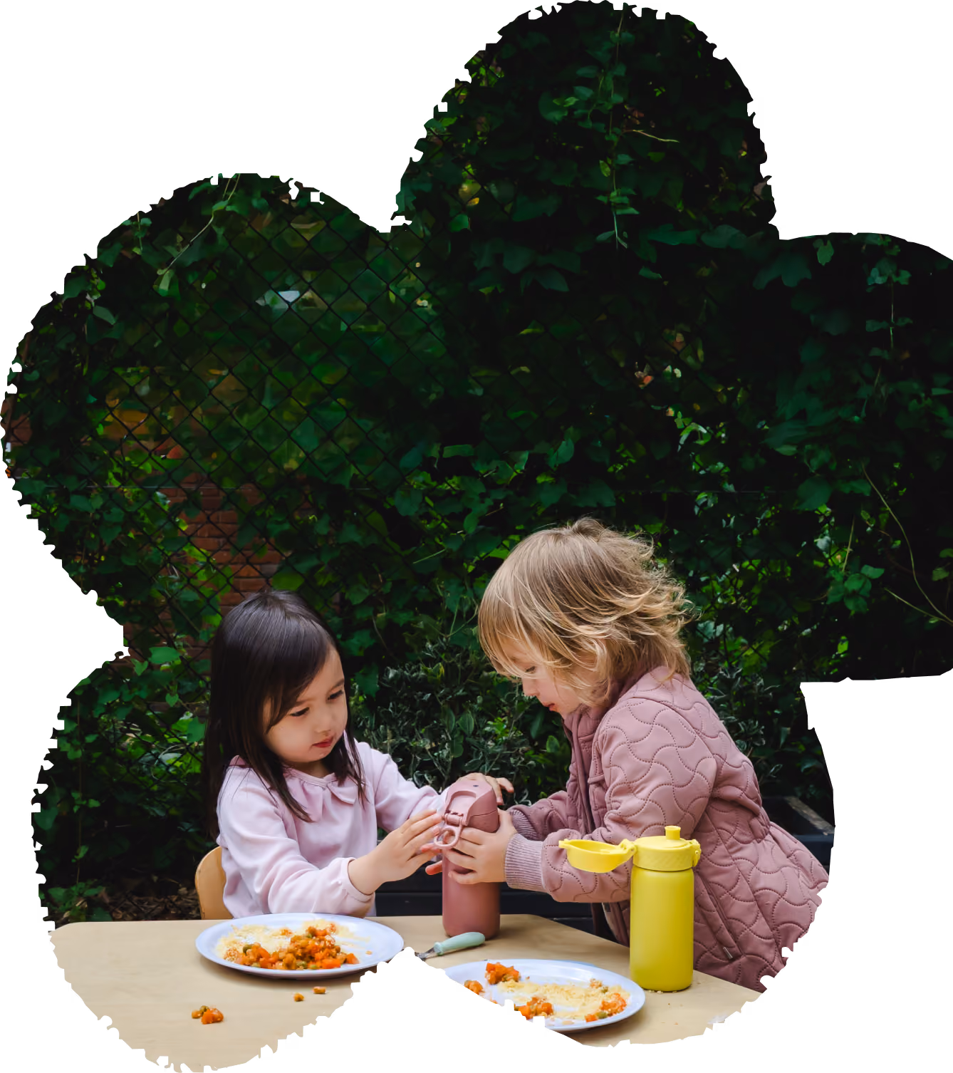 Two young children sharing a drink bottle at a table with plates of food outdoors by green foliage.