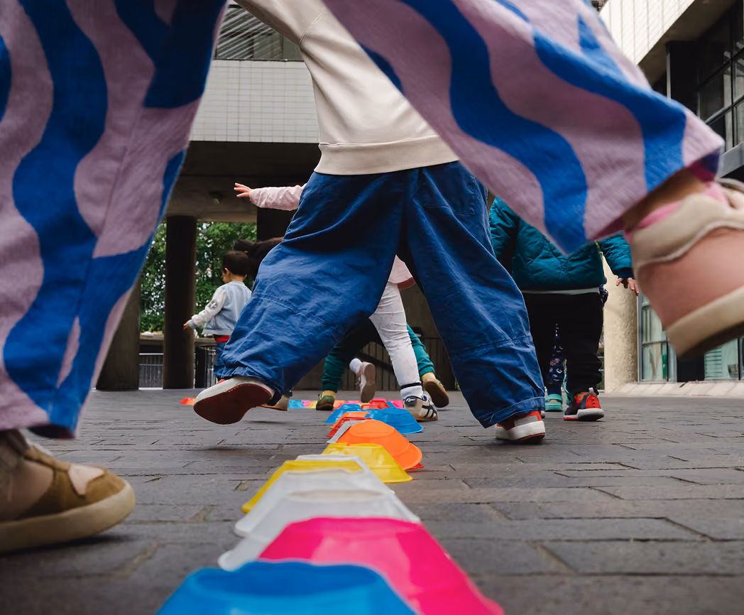 Children stepping or jumping over colorful plastic cones arranged on a paved ground outdoors.