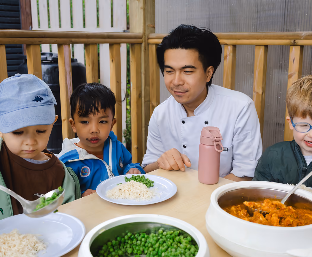 Chef serving peas and rice to three young boys sitting at a table outdoors.
