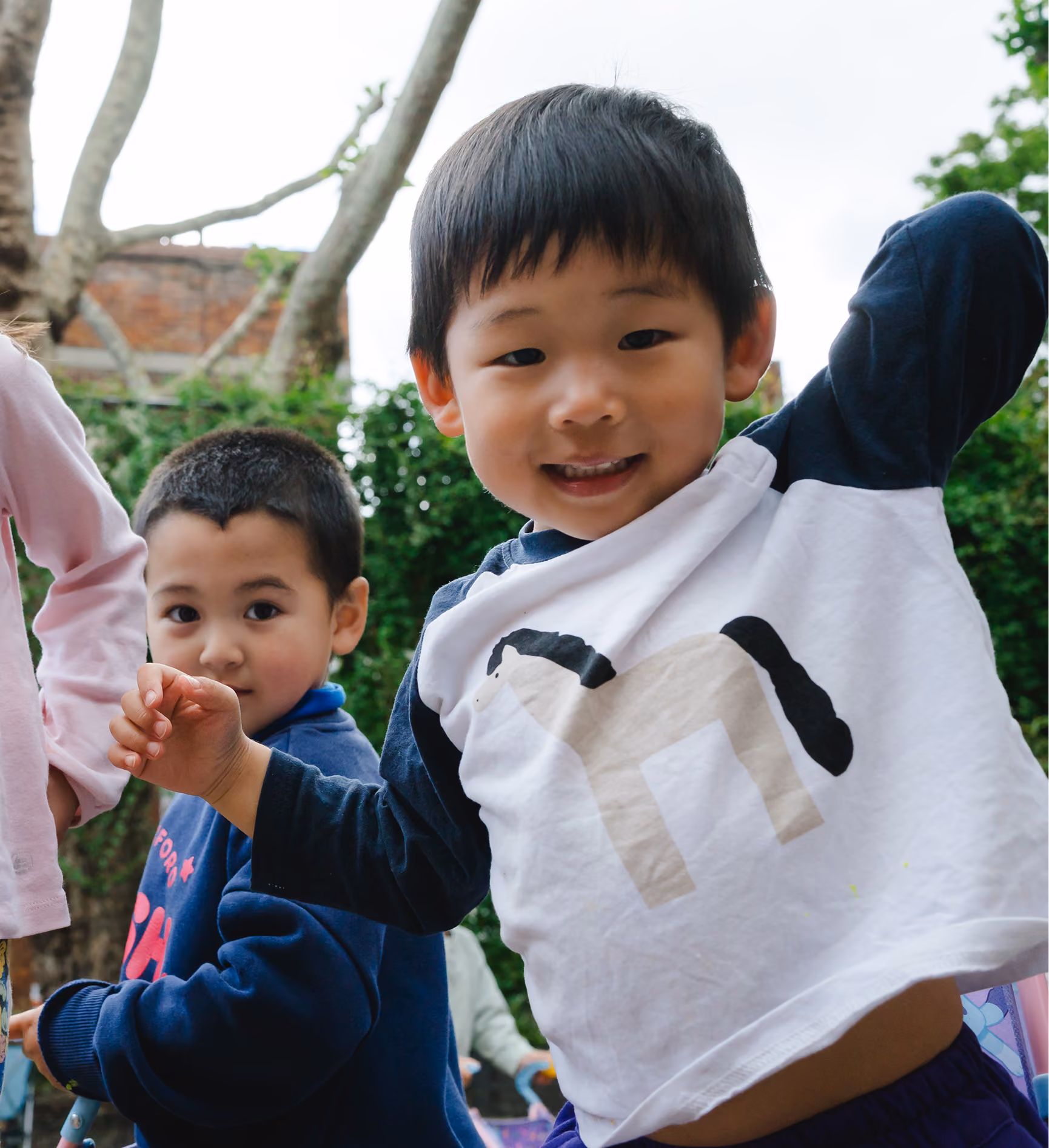 Two young boys outside playing, one smiling and moving arm, green bushes and tree in background.