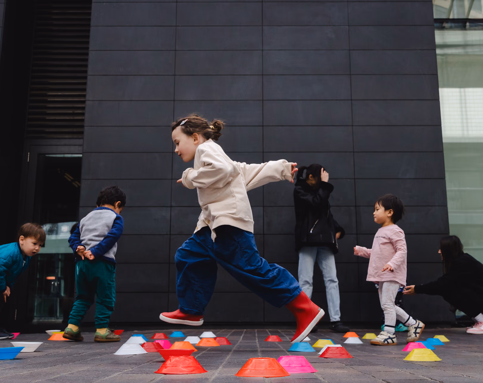 Child in beige jacket and red boots jumping over colorful cones while other kids watch on pavement.