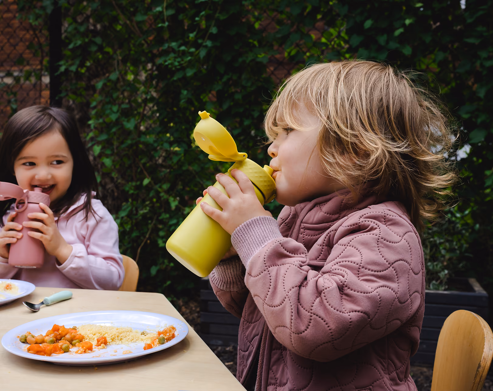 Two children sitting at a table outdoors, drinking from colorful water bottles and eating food.