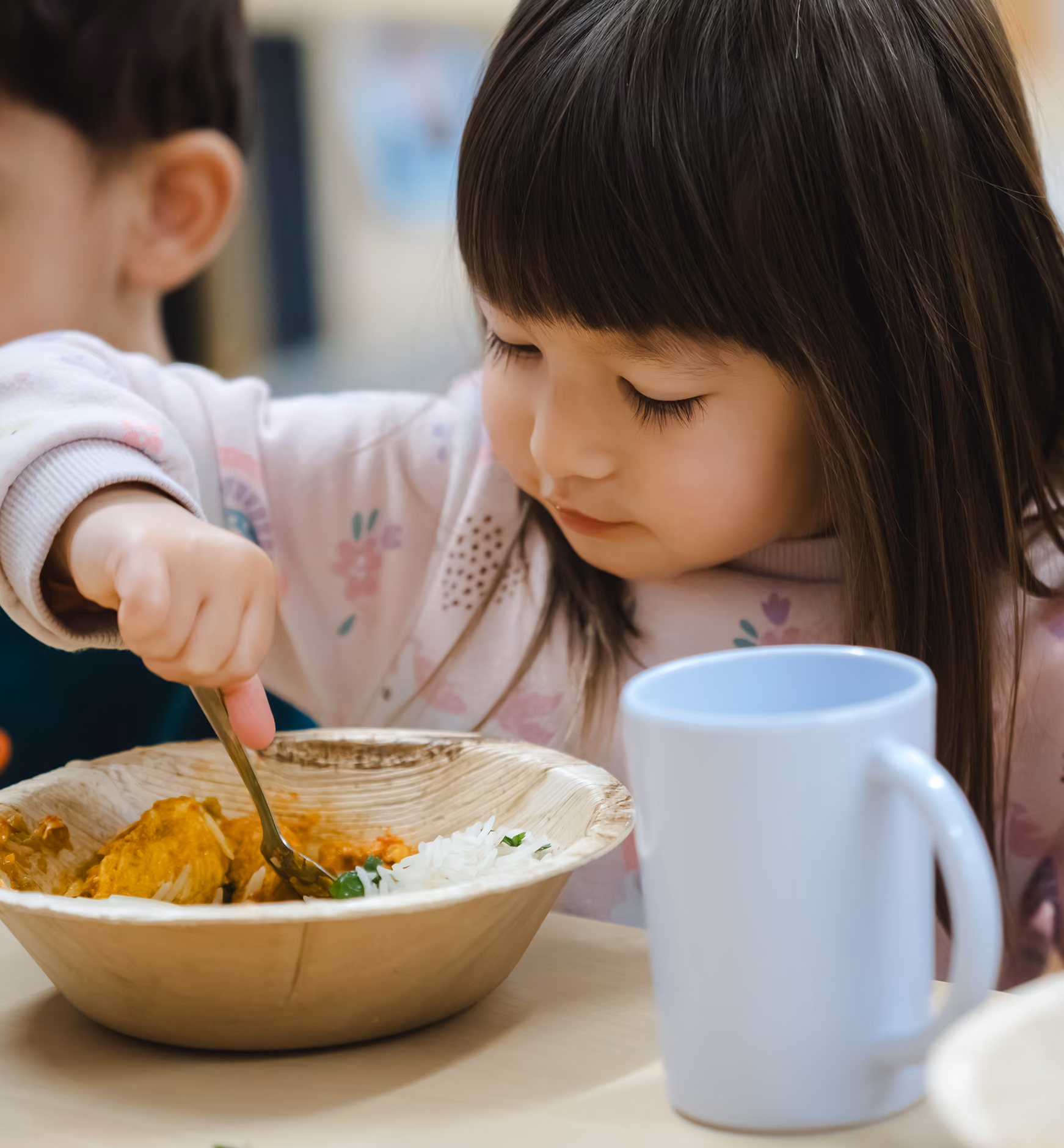 Young girl eating food with a fork from a wooden bowl next to a white cup on the table.