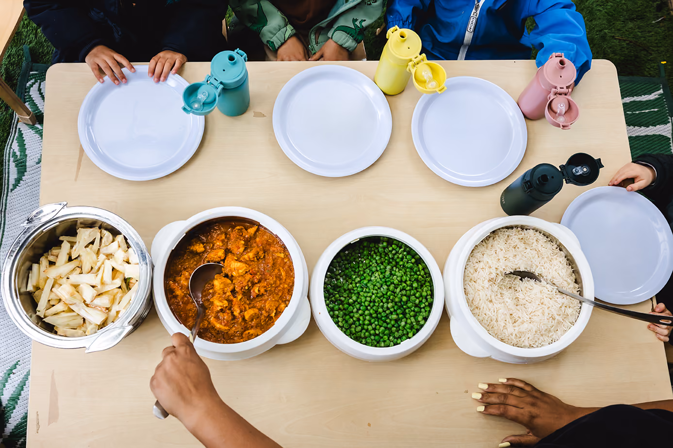 Top view of a table with empty plates, colorful bottles, and bowls of yams, curry chicken, peas, and white rice.