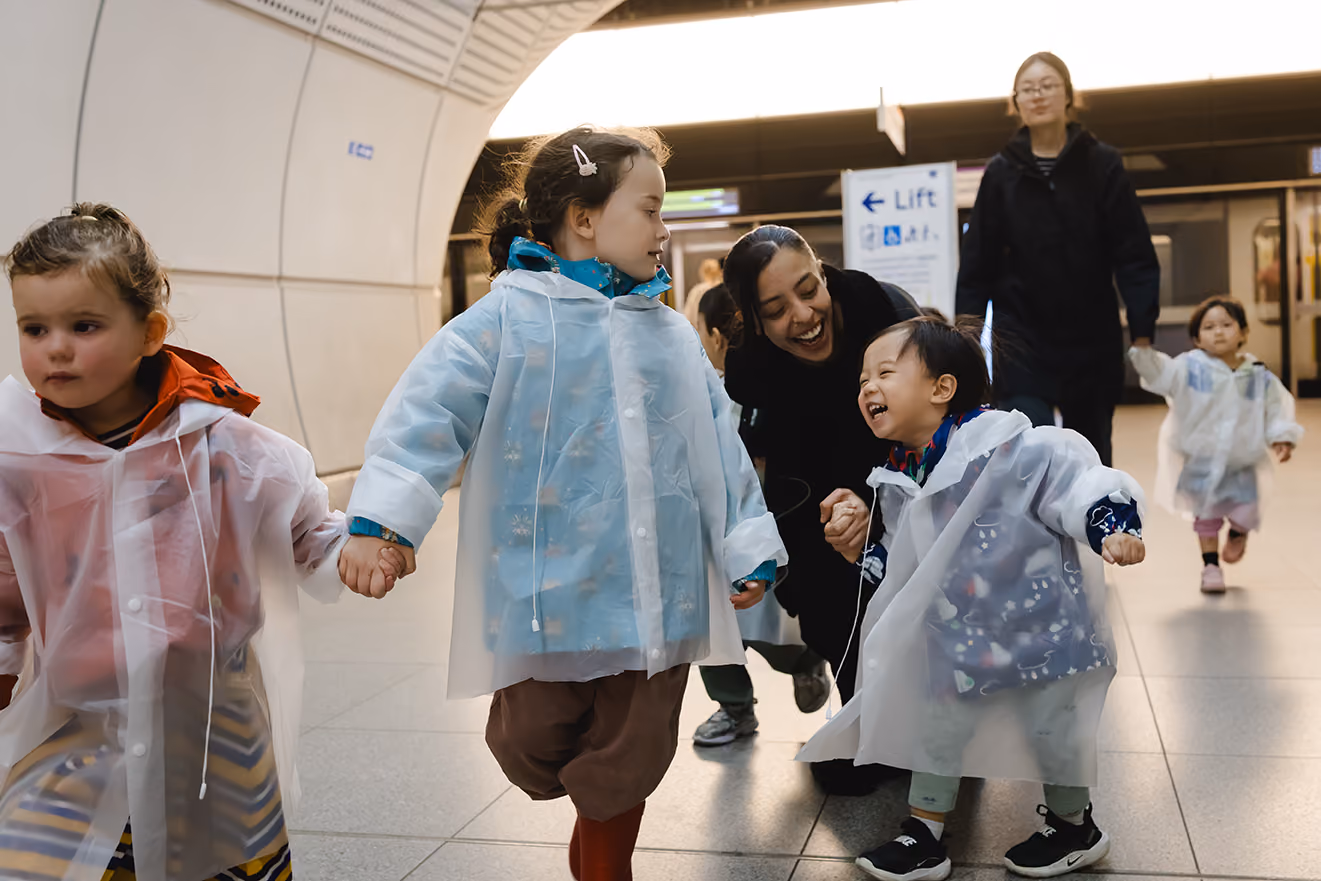 Children wearing rain ponchos hold hands and smile in a subway station with two adults nearby.