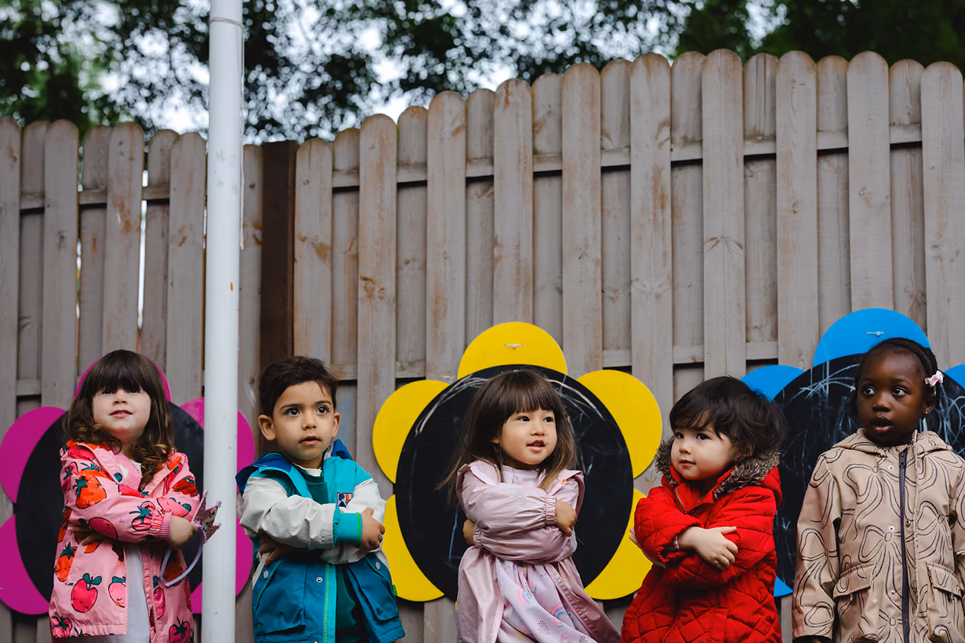 Five children in colorful jackets stand with arms crossed in front of a wooden fence with flower-shaped chalkboards.