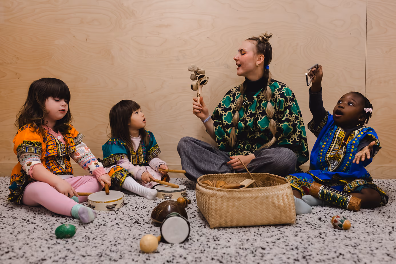 Four children in colorful African-style clothing sitting in a circle playing musical instruments.