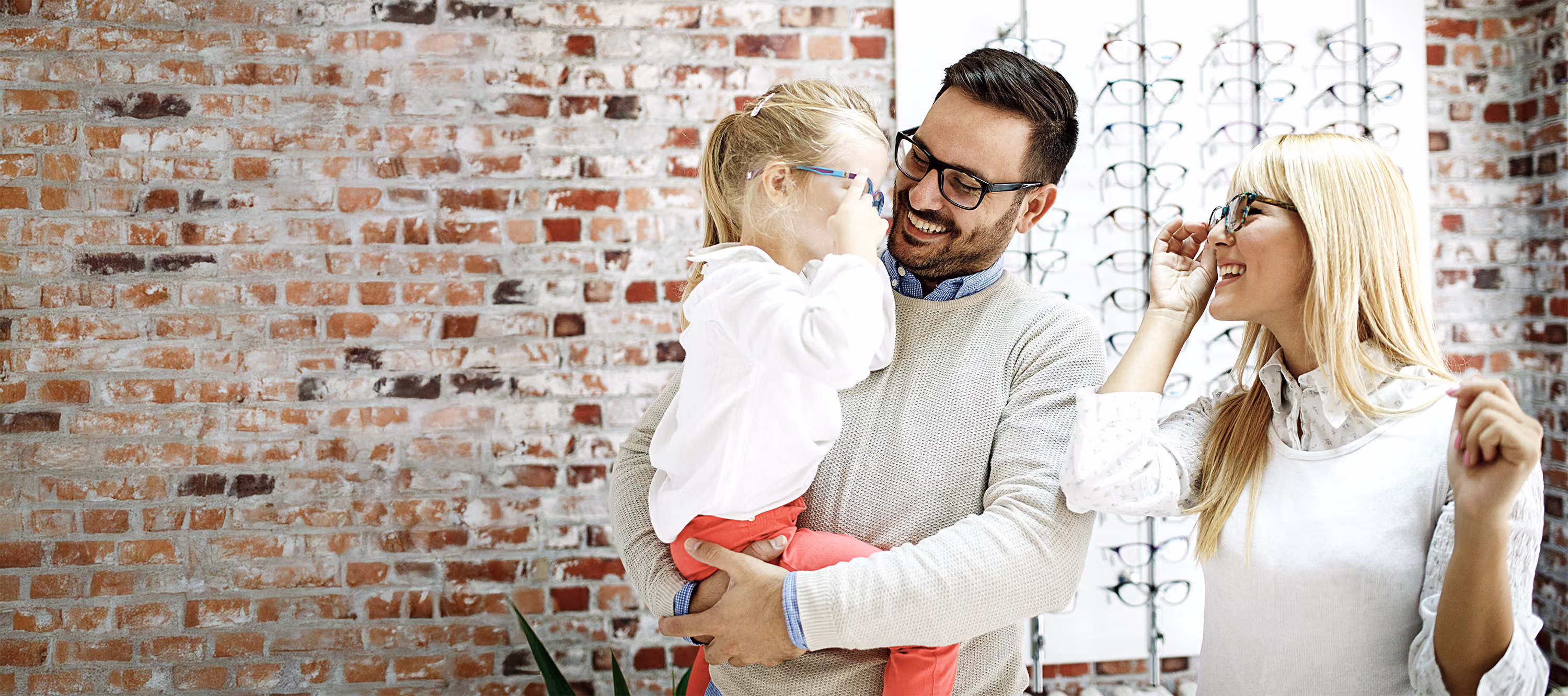 Family of three smiling and trying on eyeglasses inside an optical store with brick wall background.