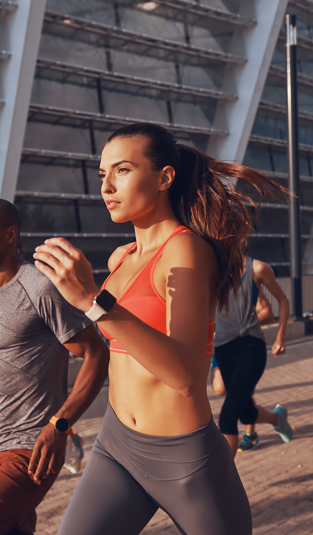 Woman in red sports bra and gray leggings running outdoors with others near a modern building.