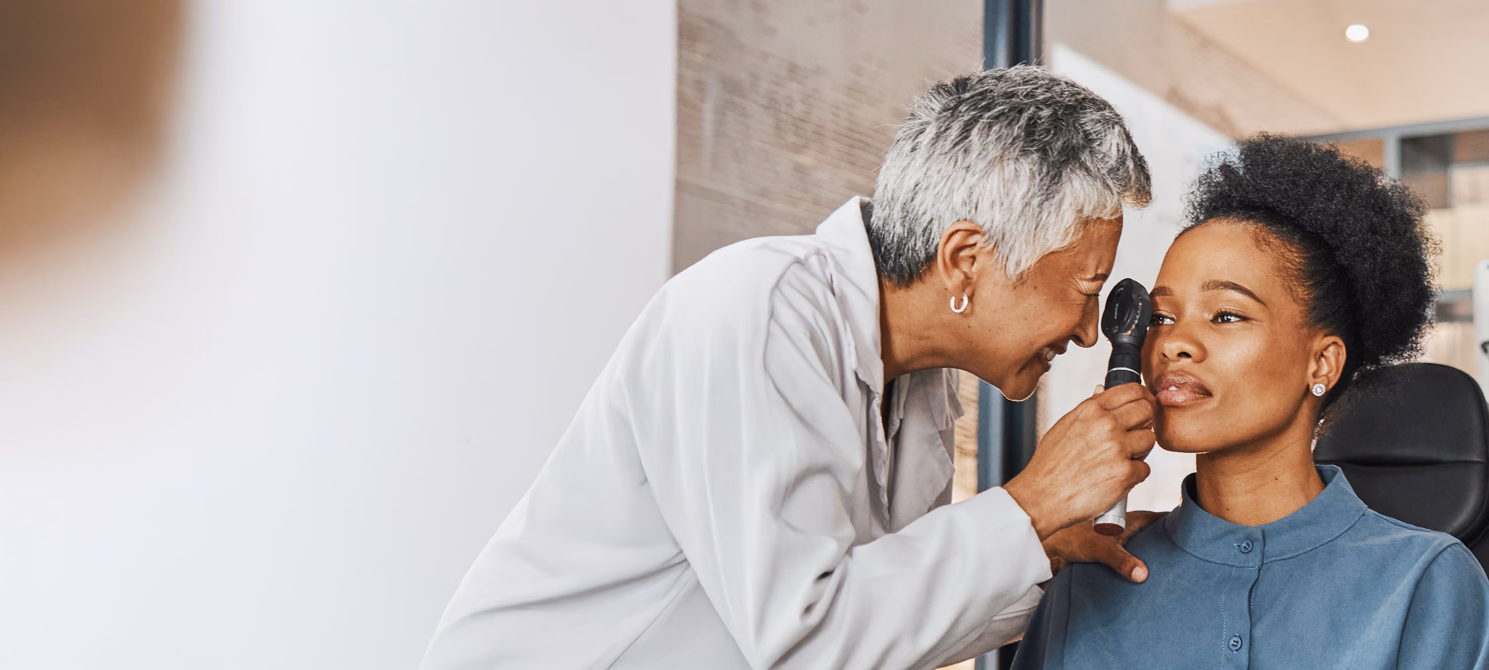 Optometrist in white coat examining a woman's eye with an ophthalmoscope during an eye exam.