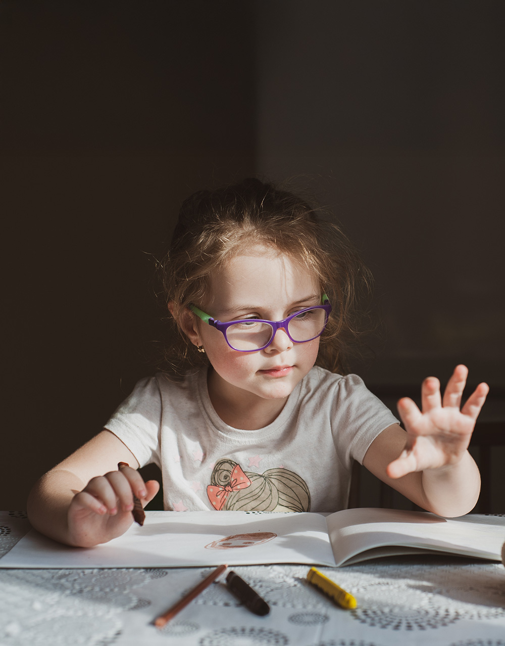 Young girl with glasses drawing with crayons in a sketchbook on a lace-covered table.
