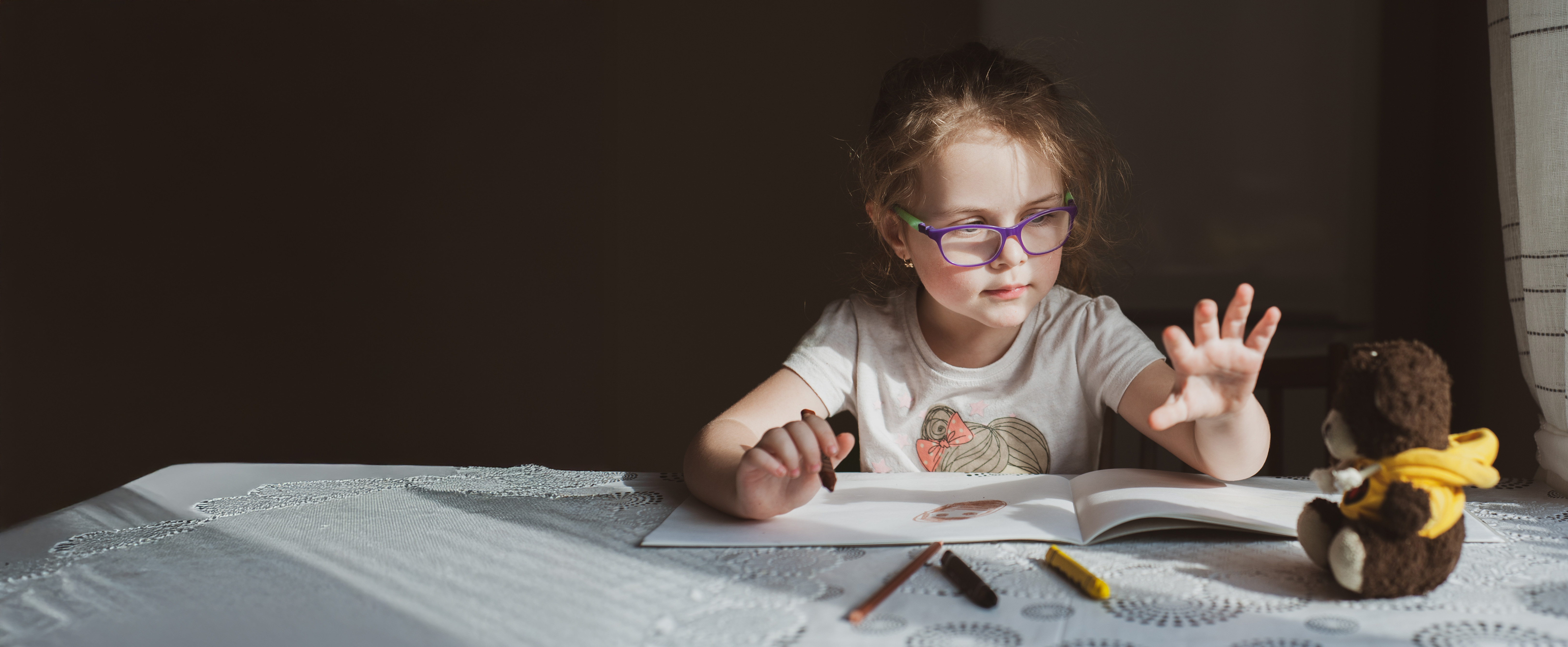 Young girl with purple glasses drawing in a book reaching toward a teddy bear on a table.