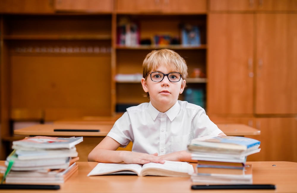 Boy wearing glasses sitting at a classroom desk with an open book and stacks of books on both sides.