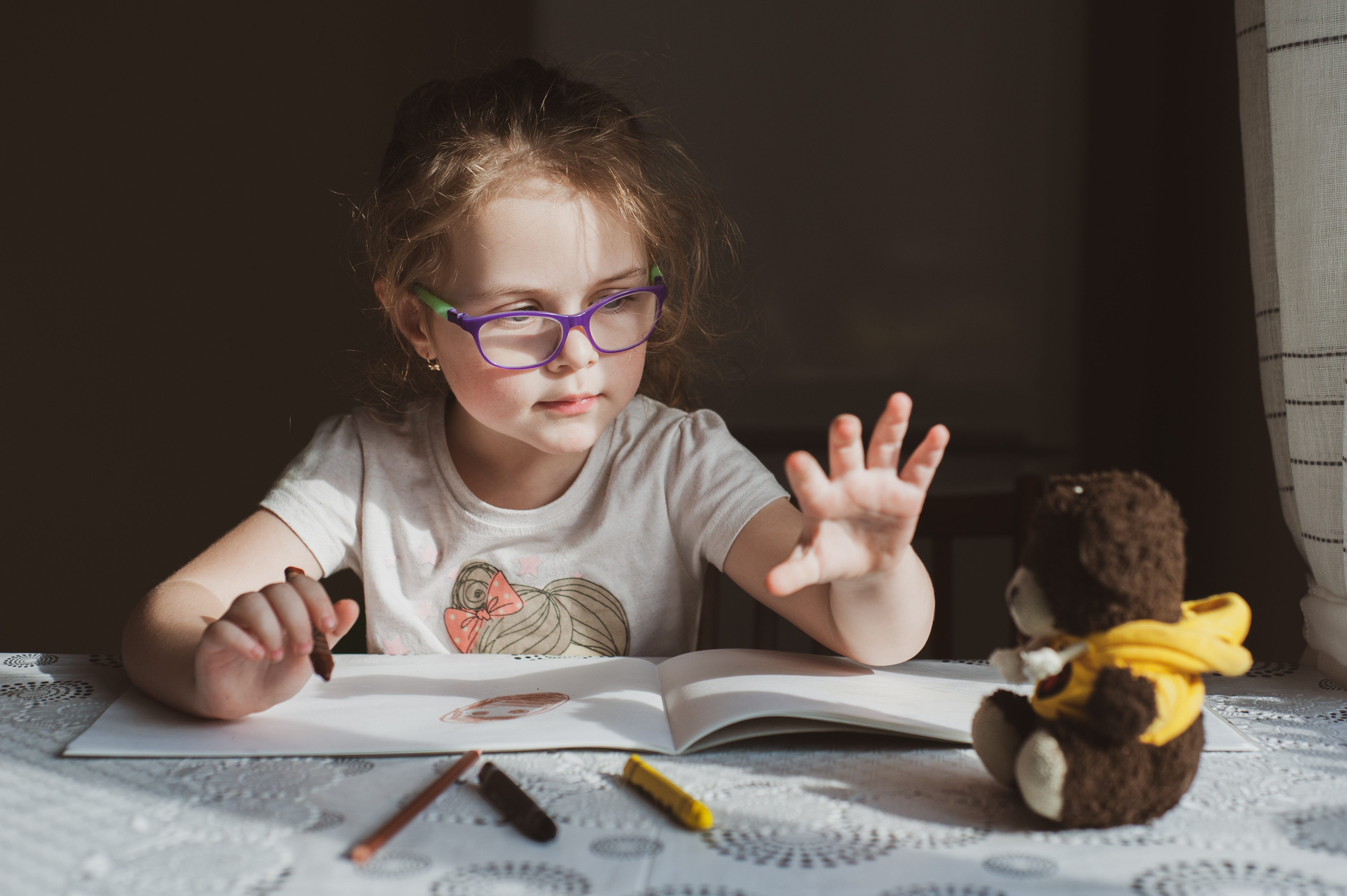 Young girl with purple glasses drawing at a table reaching out to a teddy bear in a yellow hoodie.