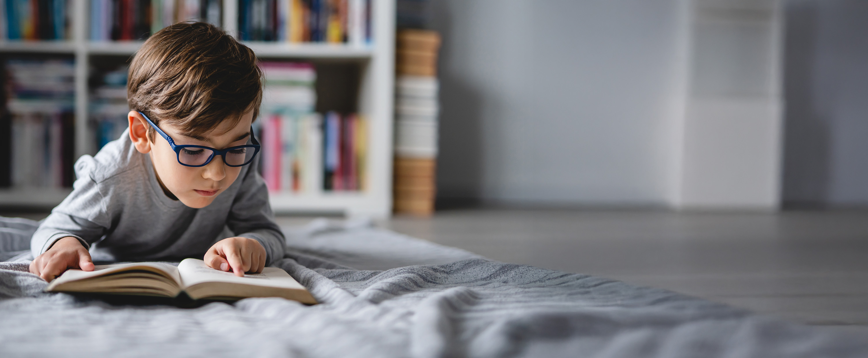 Young boy with glasses lying on a bed reading a book in a room with bookshelves in the background.