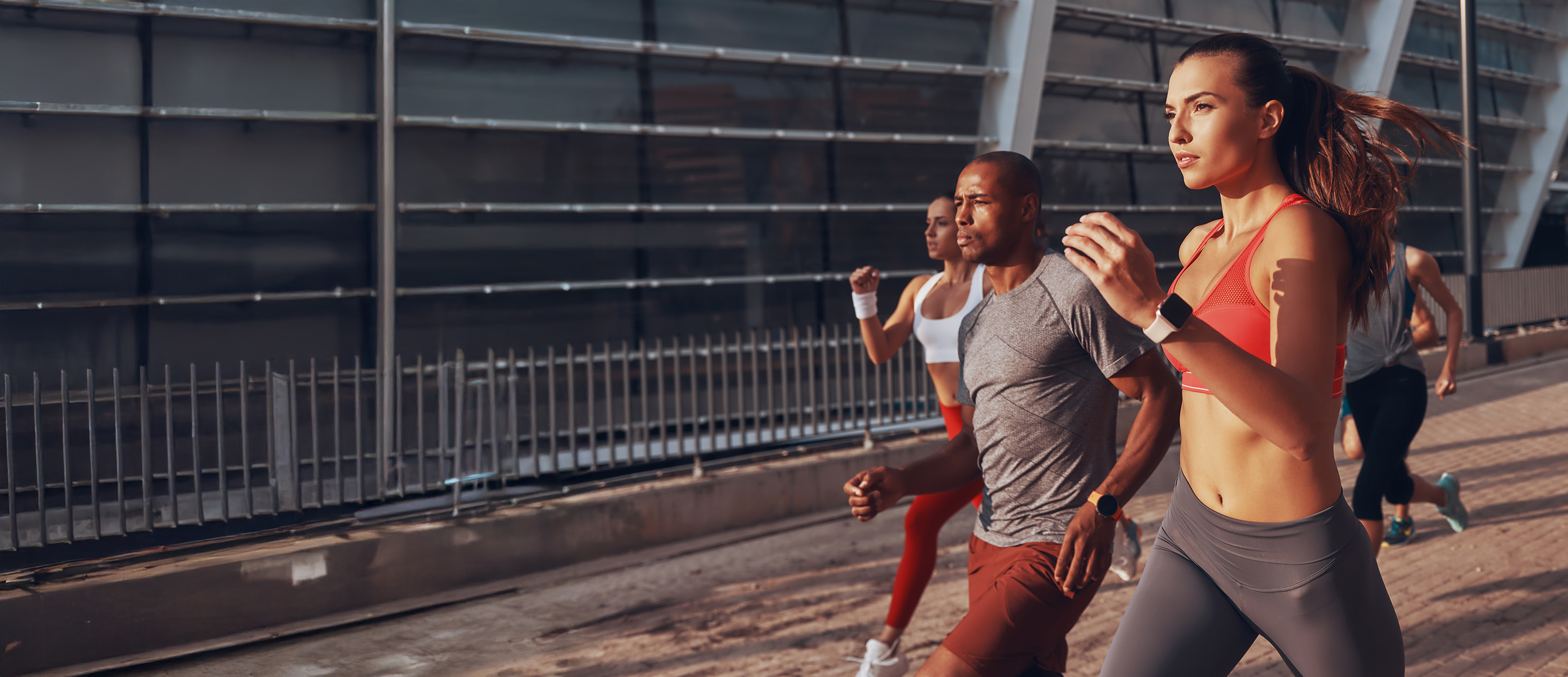 Group of fit young adults running outdoors along a pathway next to a metal fence and modern building.