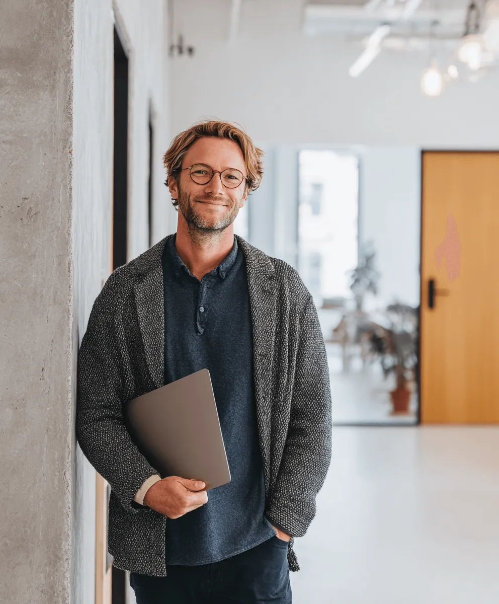 Man with glasses smiling while standing indoors holding a closed laptop.