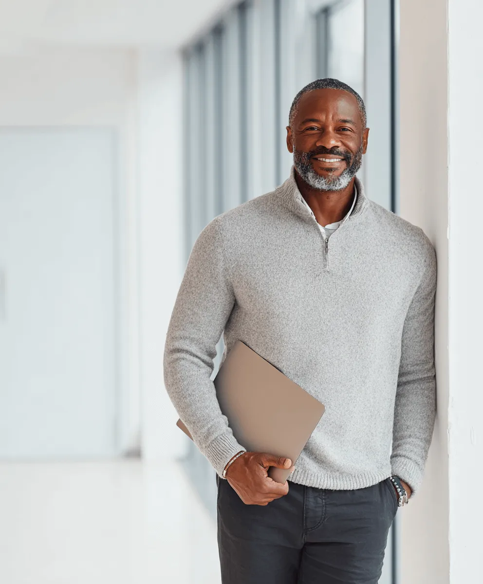 Smiling man in a grey sweater holding a laptop, standing by a window in a bright office hallway.