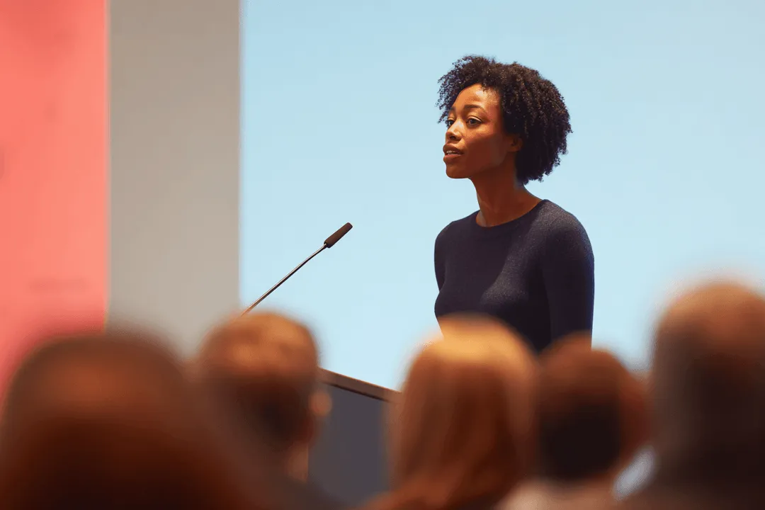 Woman with curly hair speaking confidently at a podium with a microphone in front of an audience.