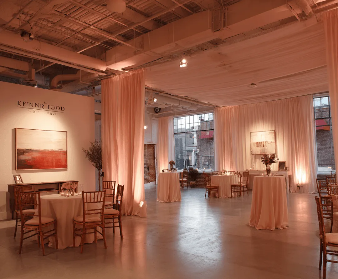 Elegant event space with round tables covered in beige cloths, wooden chairs, soft pink drapes, ambient lighting, and large windows showing a brick building outside.