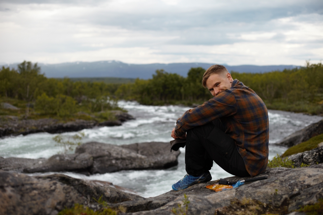 Mies istuu kalliolla kuohuvan joen rannalla tunturimaisemassa.