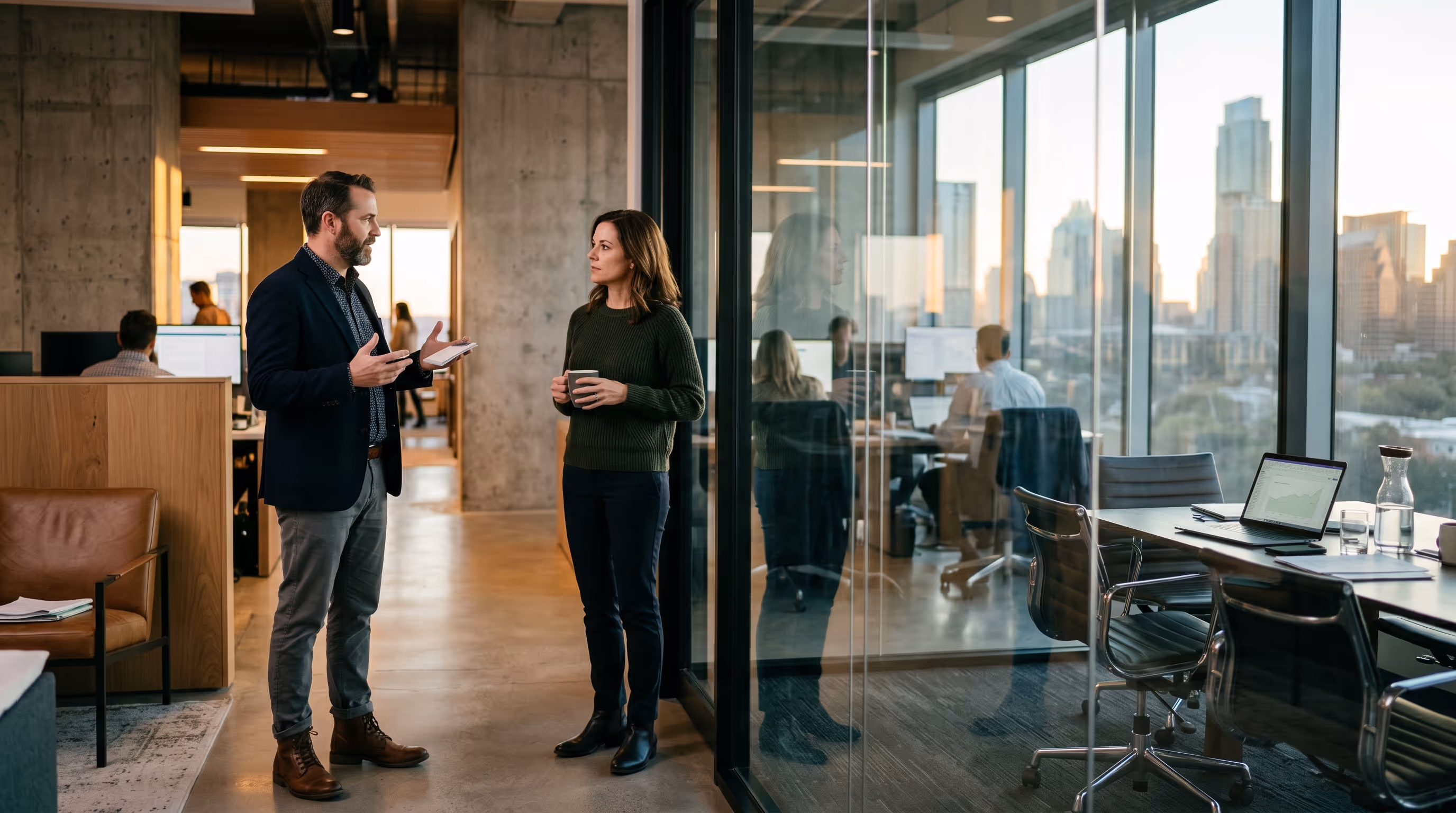 Man and woman having a discussion in an office corridor near glass-walled meeting room.