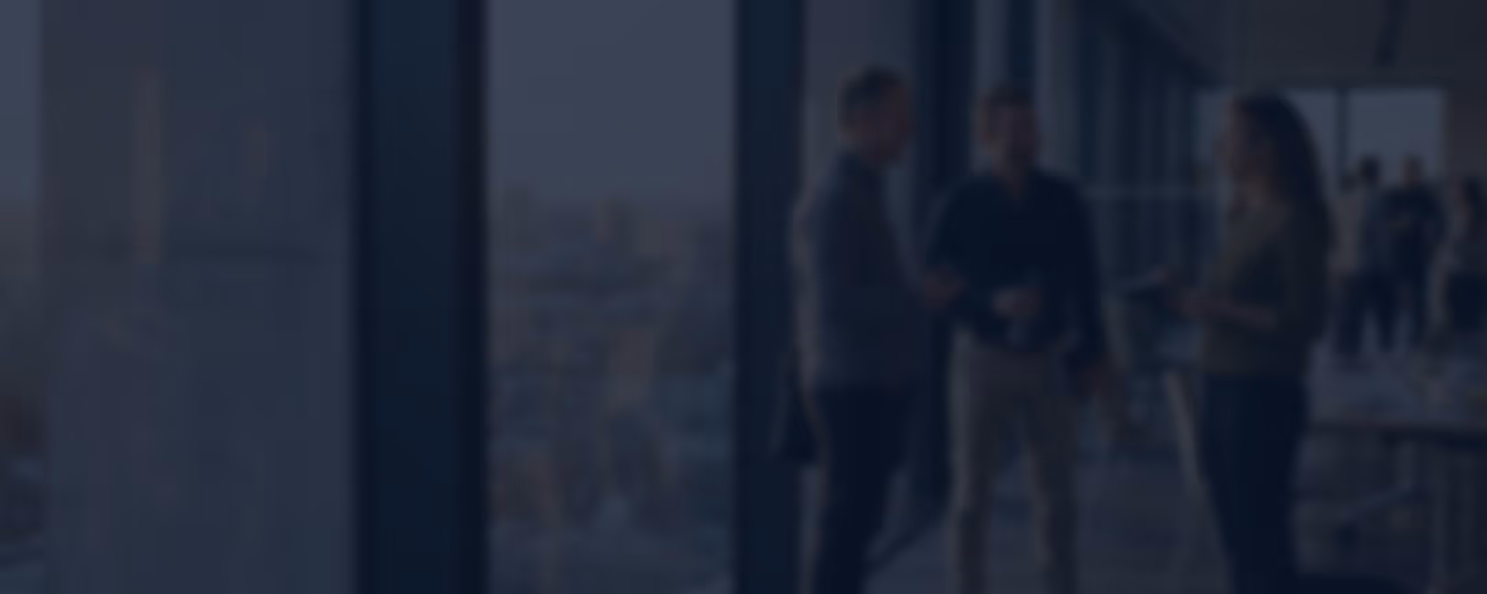 Blurry image of three people standing and talking near large windows in an office or event space.