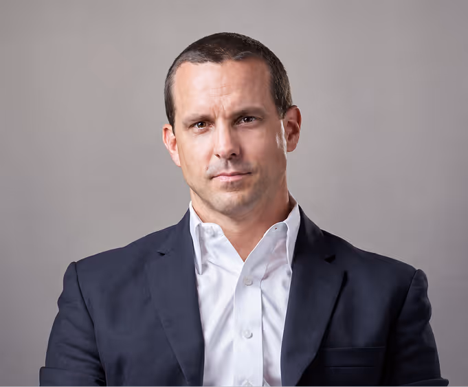 Professional man in a dark suit jacket and white shirt against a plain gray background.