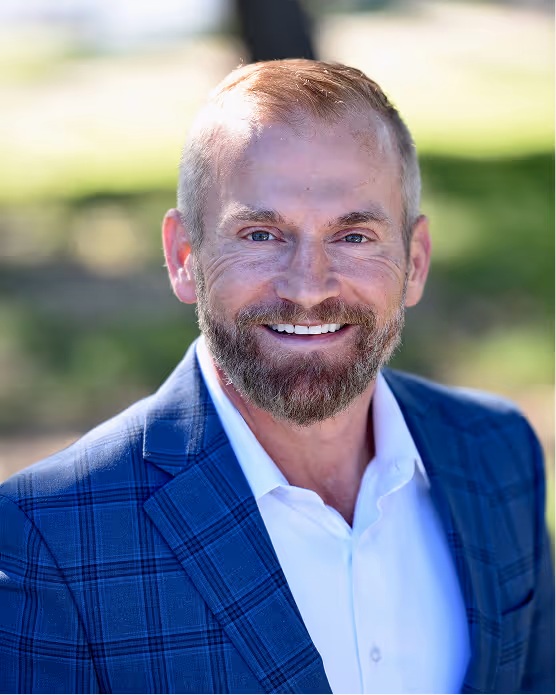 Smiling man with beard wearing a blue plaid blazer and white shirt outdoors.