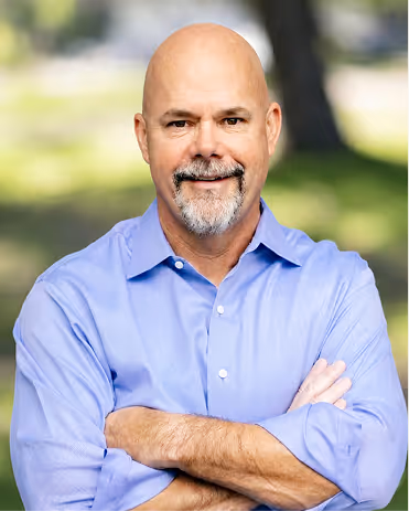 Bald middle-aged man with a gray beard wearing a light blue shirt, smiling with arms crossed outdoors.