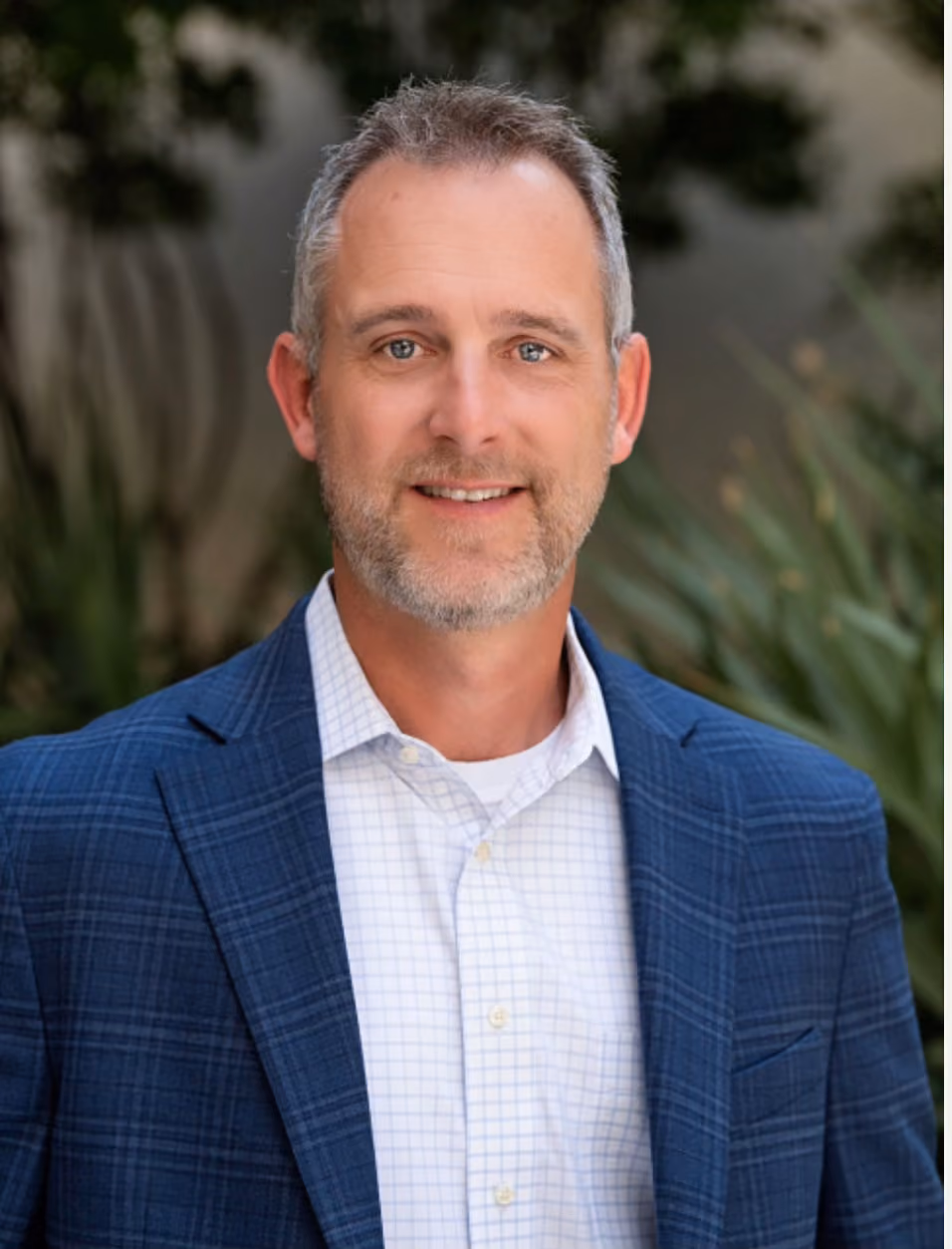Smiling man with gray hair and beard wearing blue plaid suit jacket and white checkered shirt.