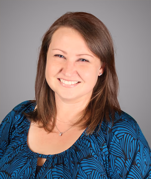 Smiling woman with shoulder-length brown hair in a blue patterned blouse on a gray background.