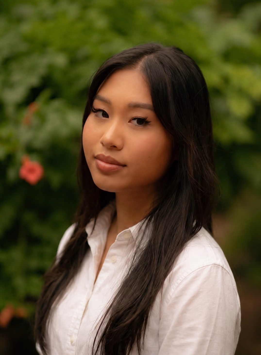 Portrait of a young Asian woman with long dark hair wearing a white shirt, standing outdoors.