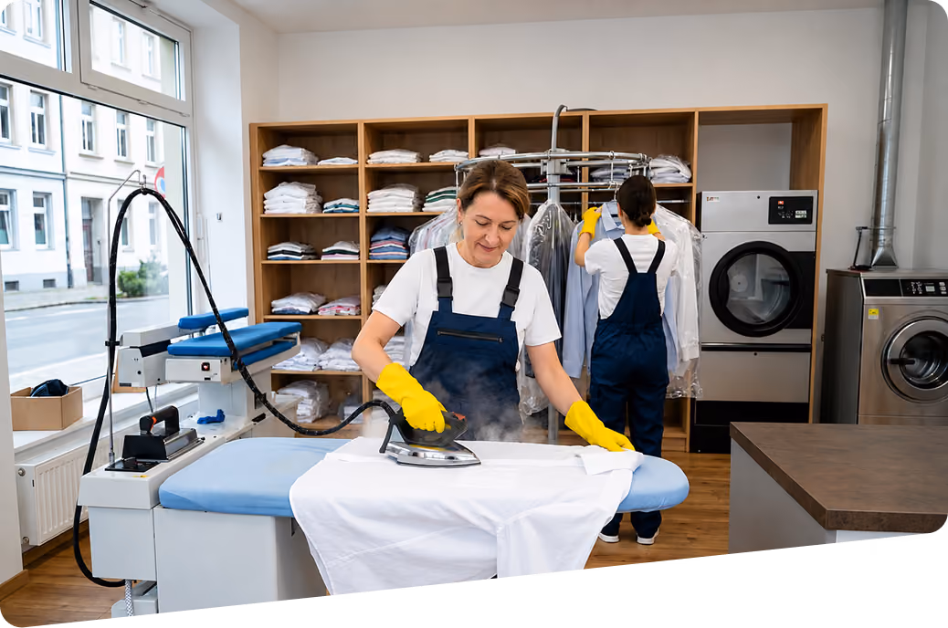 Bügelservice und Bügelhilfe: Zwei Mitarbeiterinnen in Firmenkleidung arbeiten in einer Wäscherei: Eine bügelt ein weißes Hemd, die andere hängt ein blaues Hemd nach dem Bügeln auf, während ordentlich gefaltete und gebügelte Kleidung in den Regalen liegt.