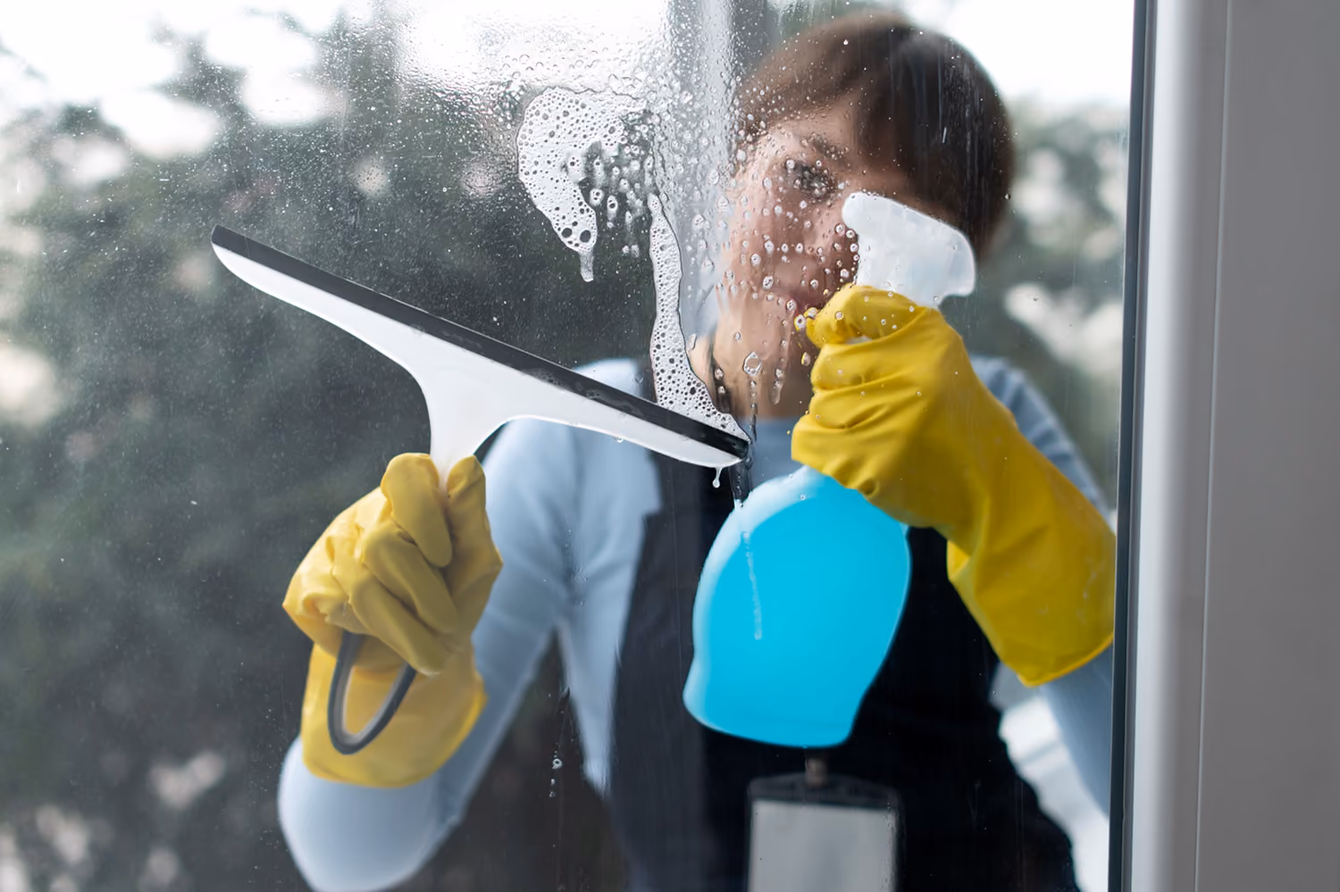 Fenster- und Glasreinigung: Reinigungskraft in Firmenkleidung reinigt ein Fenster mit einem speziellen Glasschaber und einer Sprühflasche mit Reinigungsmittel.