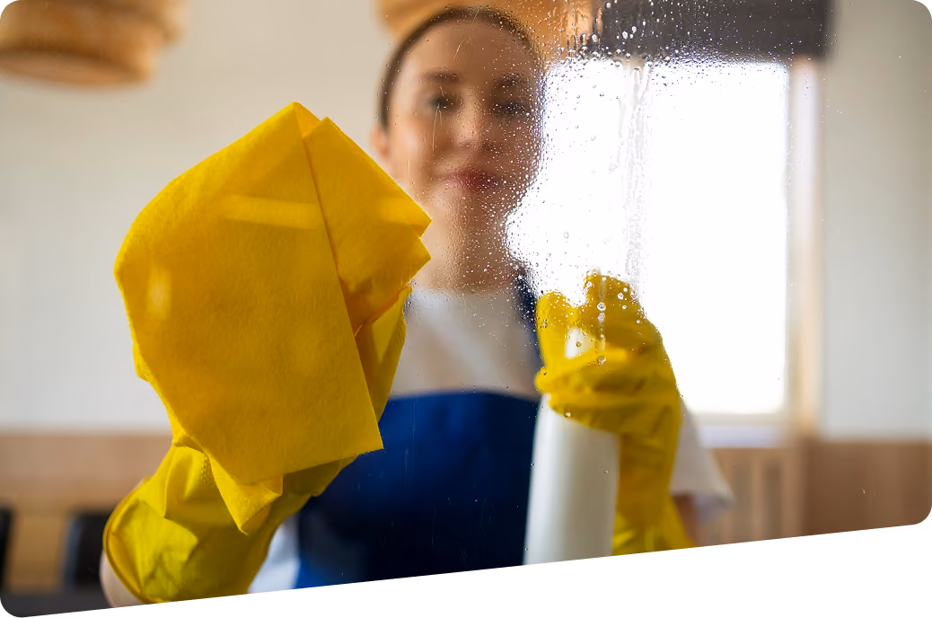 Grundreinigung: Reinigungskraft in Firmenkleidung reinigt eine Glaswand mit einem gelben Tuch und einer Sprühflasche mit Glasreiniger.