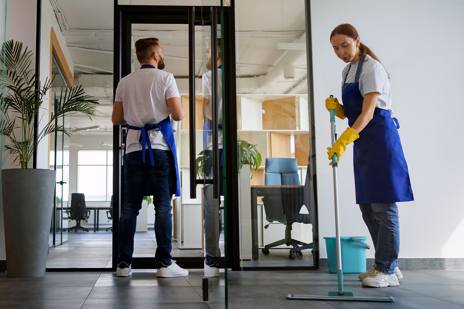 Individuelle Reinigungskonzepte für Unternehmen: Zwei Reinigungskräfte in Firmenkleidung reinigen ein Büro: Die Mitarbeiterin wischt den Boden mit einem Wischmopp, der Mitarbeiter reinigt eine Glaswand.