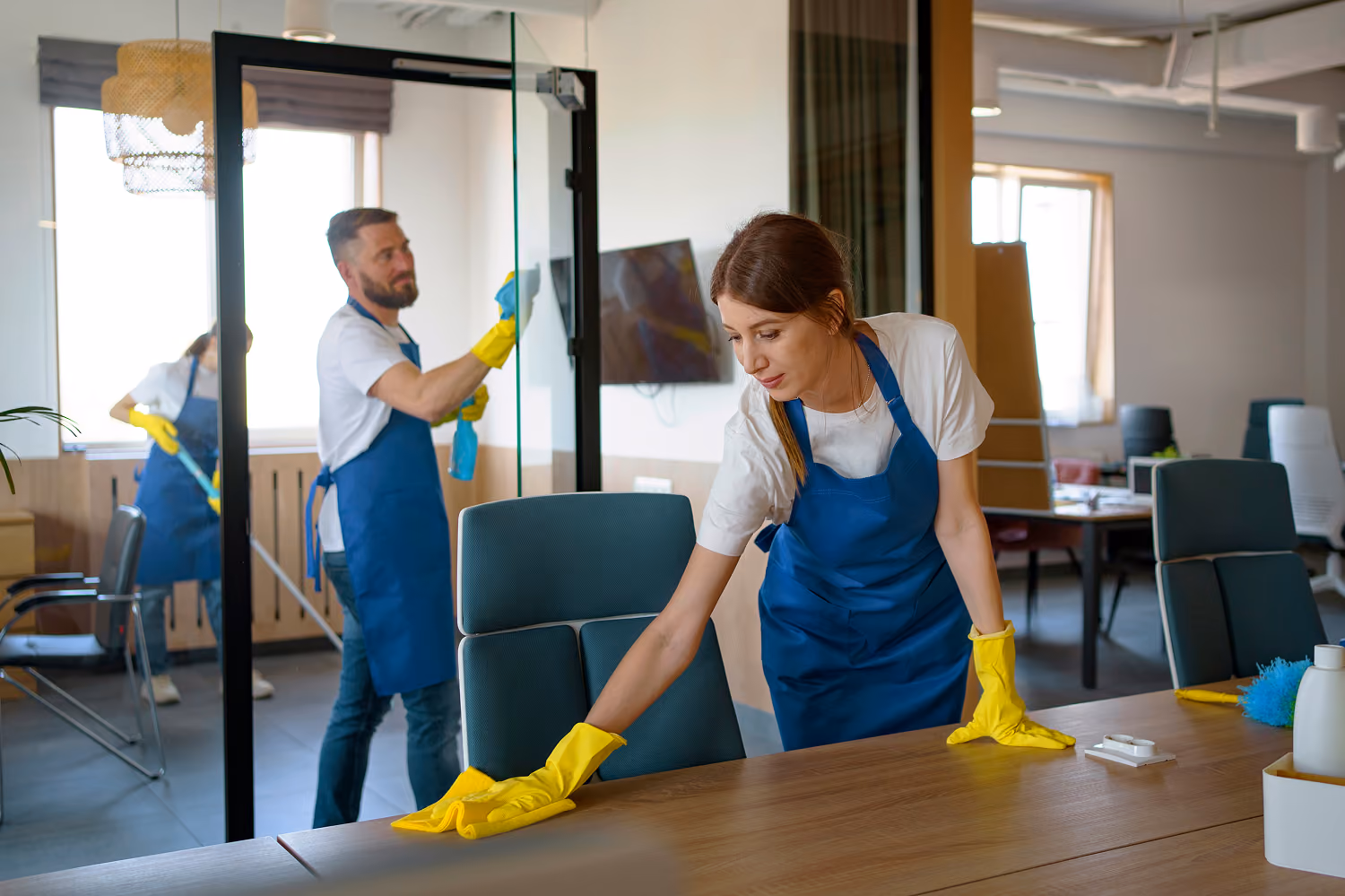 Individuelle Reinigungskonzepte für Unternehmen: Drei Reinigungskräfte in Firmenkleidung reinigen ein Büro: Eine Mitarbeiterin wischt Staub von einem großen Schreibtisch, ein Mitarbeiter reinigt eine Glastür mit Reinigungsmittel und einem Tuch, die dritte Mitarbeiterin wischt den Boden mit einem Wischmopp.