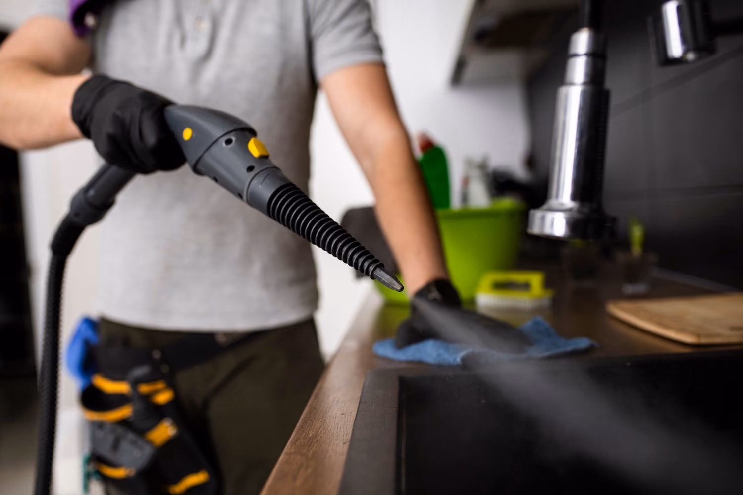 Grund- und Sonderreinigung: Reinigungskraft in Arbeitskleidung reinigt eine Kochfläche in der Küche mit einem speziellen Gerät.