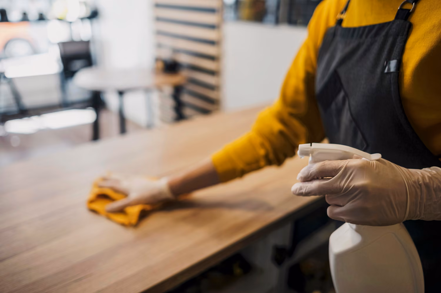 Ladenlokalreinigung: Reinigungskraft in Arbeitskleidung desinfiziert eine Arbeitsfläche in einem Café mit einem Desinfektionsmittel und einem Tuch.