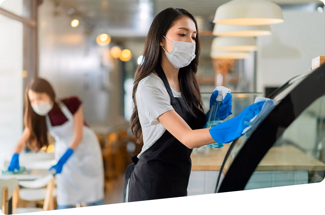 Gastronomiereinigung: Zwei Reinigungskräfte in Arbeitskleidung reinigen einen gastronomischen Bereich: Eine Mitarbeiterin reinigt eine gebogene Glasvitrine mit ausgestellten Waren, die zweite wischt einen Tisch für Gäste.